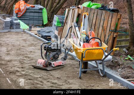 Una carriola da costruzione è riempita con vari attrezzi in un cantiere vicino a una piastra vibrante a benzina e a pile di lastre di pavimentazione. Foto Stock