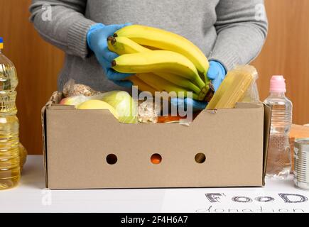 donna in guanti continua a raccogliere cibo, frutta e cose e una scatola di cartone per aiutare coloro che hanno bisogno, il concetto di aiuto e volontariato Foto Stock