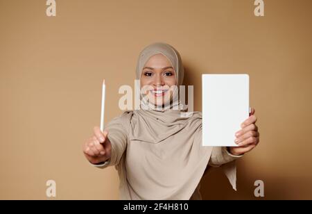 Affascinante donna musulmana araba con un bel sorriso che tiene un diario bianco e una matita. Donna d'affari in abbigliamento religioso tradizionale stretto beige b Foto Stock