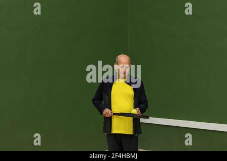 Sportivo anziano con palla da tennis e racchetta che guarda giù mentre in piedi contro la parete verde durante l'allenamento in palestra Foto Stock
