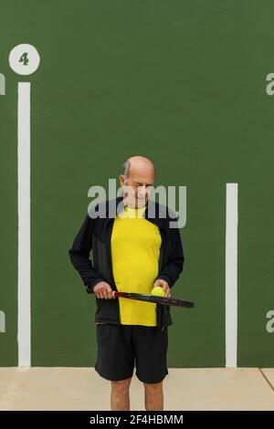 Sportivo anziano con palla da tennis e racchetta che guarda giù mentre in piedi contro la parete verde durante l'allenamento in palestra Foto Stock