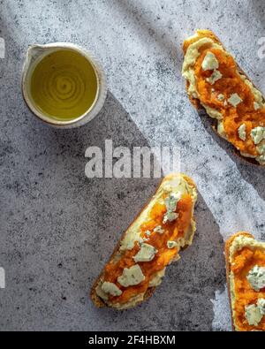 Disposizione superiore di pane di zucca sano toast con marmellata dolce e formaggio di capra messo su tavola di marmo vicino a caraffa di olio d'oliva Foto Stock