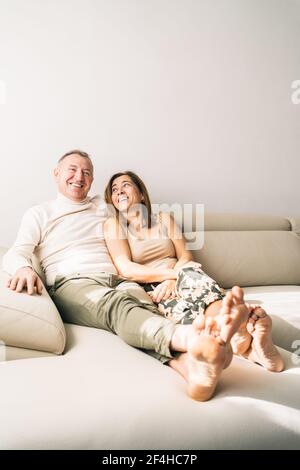 Felice coppia di mezza età seduta su un comodo divano nel soggiorno camera a casa mentre sorridi e ti godi il weekend insieme Foto Stock