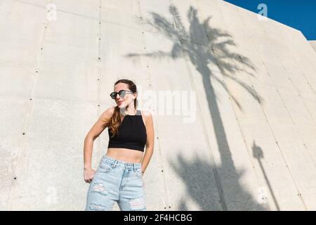 Basso angolo di elegante giovane femmina in occhiali da sole alla moda crop top nero e jeans in piedi vicino a un edificio moderno con ombra di palme tropicali Foto Stock