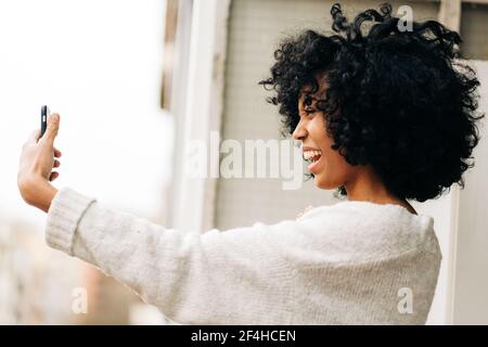 Vista laterale della carismatica femmina nera con capelli afro in piedi sul balcone e scattare da solo con lo smartphone Foto Stock