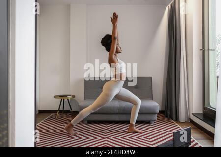 Corpo pieno di giovane femmina nera concentrata in activewear watching Video su computer portatile e Virabhadrasana esecuzione posa durante lo yoga a distanza formazione a. Foto Stock