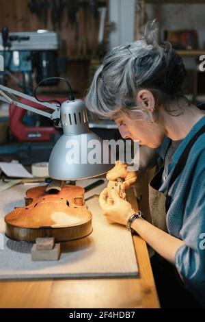 Vista laterale della decorazione artigianale in legno da taglio femminile per la lavorazione a mano violino durante il lavoro in officina Foto Stock