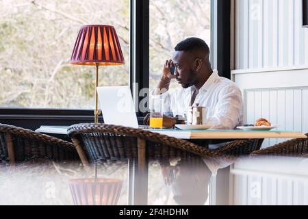 Serio uomo d'affari africano americano adulto seduto al tavolo con il computer portatile e discutere di problemi aziendali durante la telefonata mentre si ha la colazione in un ambiente accogliente Foto Stock