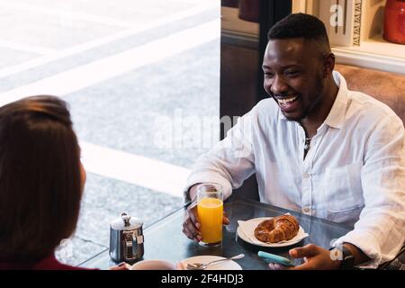 Coppia multietnica che ha una conversazione felice mentre si siede al tavolo vicino finestra e fare colazione insieme in un elegante e accogliente caffè Foto Stock