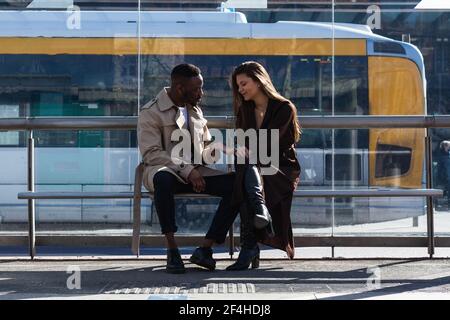 Corpo pieno di elegante coppia multietnica amorevole in abiti eleganti tenere le mani e parlare mentre si è seduti sulla panchina dell'autobus stazione in città Foto Stock