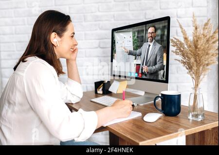 Formazione online. Videochiamata. Giovane donna soddisfatta, studiando a distanza, prendendo appunti, sullo schermo del computer il tutor maschile conduce una lezione online che mostra informazioni su una lavagna a fogli mobili, studiando da casa Foto Stock