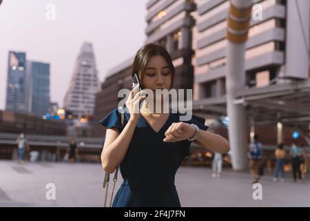 Giovane donna asiatica di successo in abbigliamento casual check-in tempo orologio da polso in attesa di qualcuno e di parlare al telefono strada cittadina di notte Foto Stock