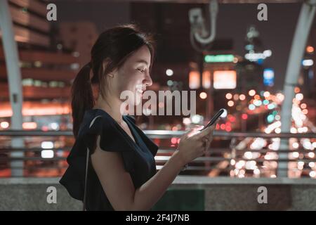Vista laterale della bella giovane donna asiatica felice che si erge sopra il ponte pedonale controllare lo smartphone e navigare tra le applicazioni durante il controllo della posta elettronica Foto Stock
