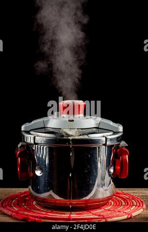 Un fornello a pressione in acciaio si sta raffreddando su un trivet di tessuto su un tavolo di legno. La valvola di pressione viene rilasciata e il vapore fuoriesce quando il sistema si depre Foto Stock