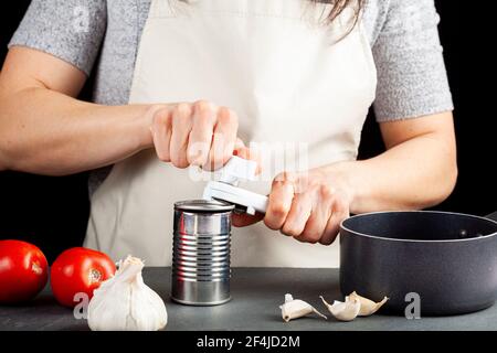 Una donna sta aprendo con attenzione una lattina di pasta di pomodoro su un banco da cucina utilizzando un apriboccatole in plastica bianca. Sta preparando un pasto per cui usa b Foto Stock