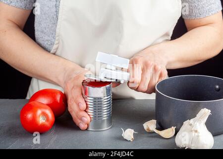 Una donna sta aprendo con attenzione una lattina di pasta di pomodoro su un banco da cucina utilizzando un apriboccatole in plastica bianca. Sta preparando un pasto per cui usa b Foto Stock