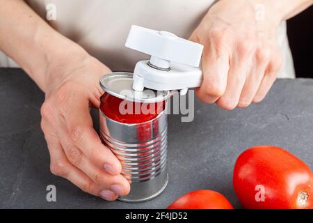 Una donna sta aprendo con attenzione una lattina di pasta di pomodoro su un banco da cucina utilizzando un apriboccatole in plastica bianca. Sta preparando un pasto per cui usa b Foto Stock