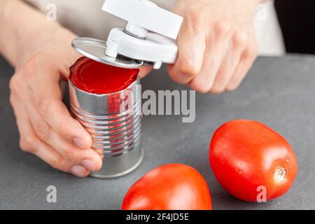 Una donna sta aprendo con attenzione una lattina di pasta di pomodoro su un banco da cucina utilizzando un apriboccatole in plastica bianca. Sta preparando un pasto per cui usa b Foto Stock