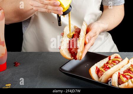 Una donna caucasica sta preparando panini caldi del cane. Sta spremendo la mistrd dalla bottiglia di plastica sopra ad una salsiccia di manzo all'interno di un mordere caldo del cane. Altri sandw Foto Stock