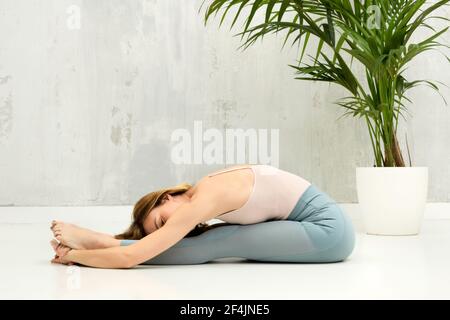 Donna che fa una Pascimottanasana o seduta Forward Bend yoga posa per rilassarsi e allungare le gambe e la colonna vertebrale con copyspace su una parete grigia oltre Foto Stock