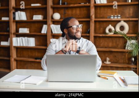 Felice premuroso giovane afroamericano in occhiali seduti alla scrivania in ufficio, guardando via, prendendo la pausa dal lavoro sul computer portatile, pensando a nuove idee, sorridendo e pianificazione fine settimana o vacanza Foto Stock