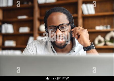 Giovane operaio africano americano maschio in occhiali e cuffie rispondere alla chiamata, guardando lo schermo del computer portatile e ascoltando attivamente le richieste nel reparto di assistenza clienti, tenendo in mano il microfono Foto Stock