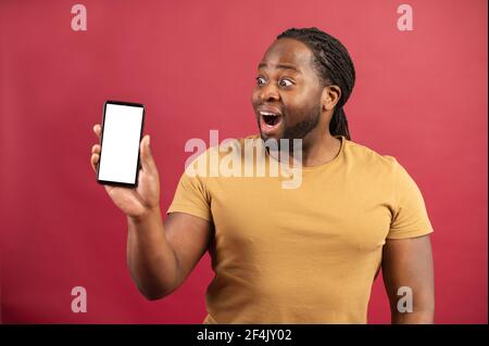 Sorpresi dalle possibilità di una nuova app mobile afro-americano che fissa lo smartphone con schermo vuoto, il sorprendente ragazzo con pelle scura dimostra il telefono con display vuoto, isolato in rosso Foto Stock