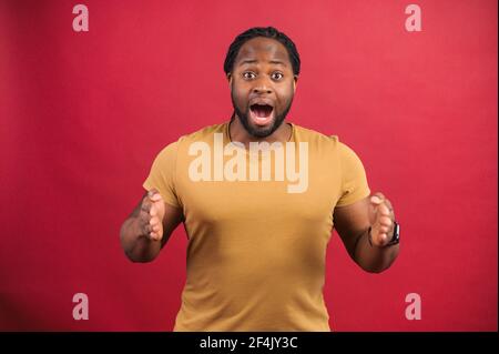 L'uomo africano sorpreso che tiene le mani davanti a lui, presentando qualcosa, scioccato ragazzo skinned scuro che tiene spazio vuoto per il vostro prodotto isolato su rosso, dimostra la dimensione. Concetto di mockup Foto Stock