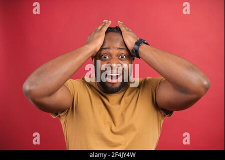 Uomo afro-americano spaventato e scioccato che regge la testa, guarda la macchina fotografica in incredulità, ragazzo azzardato dalla pelle scura con la bocca aperta isolata in rosso Foto Stock