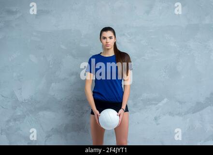 Pallavolo ragazza tenere e calcio palla in costume blu su sfondo grigio parete in cemento. Giocatore che fa allenamento sportivo a casa. Concetto di sport e ricreazione Foto Stock