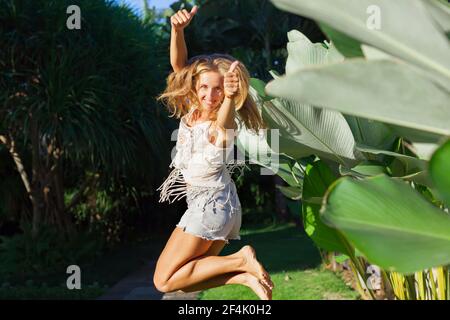 Felice giovane donna si diverte in un resort tropicale. In villa di lusso giardino divertente ragazza salto alto mostra pollici su. Stile di vita attivo, attività di viaggio delle persone Foto Stock