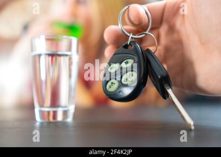 La mano dell'uomo tiene le chiavi dell'automobile in primo piano sullo sfondo di una pila di vodka e di una ragazza. Concetto di guida ubriaca Foto Stock