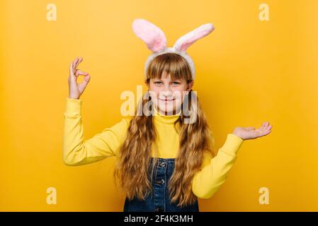 Carino adolescente caucasica ragazza in conigliato fiaccie orecchie alza palma e mostrando il segno di mano OK, pollice su come gesto, modelli su sfondo giallo studio Foto Stock