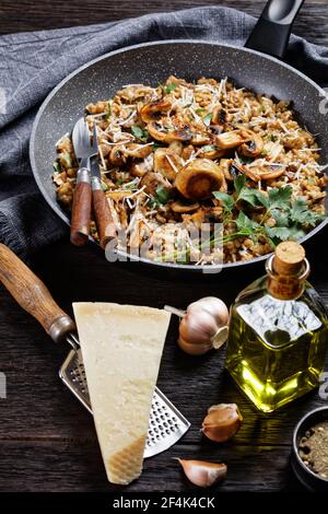 classico risotto cremoso ai funghi cosparso di parmigiano grattugiato in una padella e ingredienti su un tavolo di legno scuro, cucina italiana Foto Stock