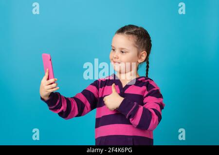 primo piano ritratto di una ragazza sorridente con pigtail che parla su una chat video del telefono e che dà un pollice in su, isolato su uno sfondo blu Foto Stock