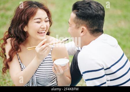 Ridendo bella giovane donna vietnamita che dà un pezzo di suchi a. ragazzo quando si godono un picnic romantico nel parco Foto Stock