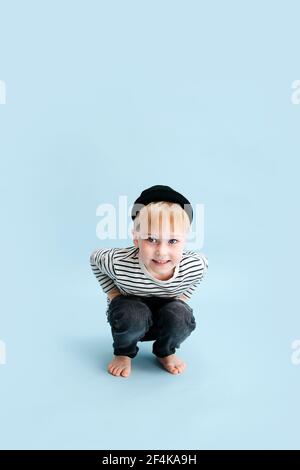 Sorridendo piccolo biondo ragazzo che squatting. Indossa una camicia a manica lunga a righe e un cappuccio nero. Scatto in studio. Su sfondo blu. Foto Stock