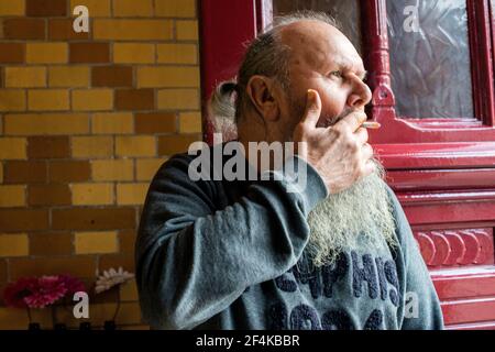 Tilburg, Paesi Bassi. Senior maschio adulto con barba di fumare un cigerette vicino alla porta di ingresso di un ristorante cafe. Foto Stock