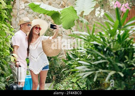 Felice giovane coppia sorridente con valigie che prendono selfie in giardino di un lussuoso resort termale Foto Stock