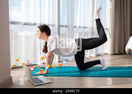 Giovane donna caucasica in abbigliamento sportivo facendo yoga live online. Interno. Emoticon ed emoticon volano intorno al laptop. Il concetto di social network, online Foto Stock