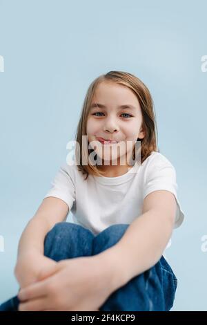 Seduta 9 anni ragazza con il sorriso perky e appena la punta della sua lingua bloccato fuori su sfondo blu. Indossa jeans blu e camicia bianca. Si sita Foto Stock