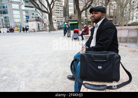 New York City, Stati Uniti. Borsa per computer portatile afroamericana maschio strega, in attesa alla panchina Columbus Circle - Centrasl Park. Foto Stock