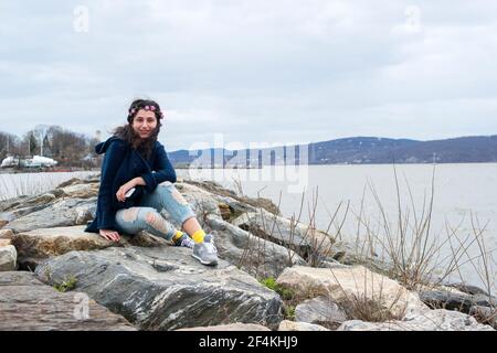 Peekskill, New York, Stati Uniti. Ritratto di un giovane adolescente gil seduto su un molo roccioso presso la Hudson River Shoreline. Foto Stock