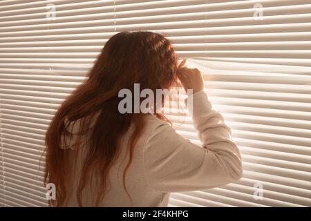 Vista posteriore di bella giovane donna con i capelli lunghi sbirciare attraverso il foro nelle tende della finestra e guarda fuori la finestra. Concetto di sorveglianza e curiosità Foto Stock