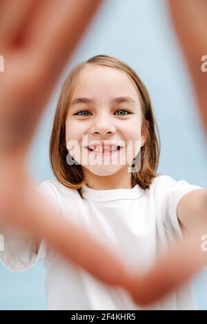 Ritratto di una ragazza sorridente di 9 anni attraverso un dito cornice che fa. Su sfondo blu. Scatto in studio. Manca un dente. Foto Stock
