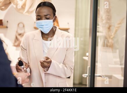 Donna afroamericana proprietario del negozio che applica l'igienizzatore a un cliente mani Foto Stock