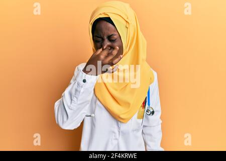 Bella giovane donna africana che indossa uniforme medico e hijab odore qualcosa di stinoso e disgustoso, odore intollerabile, trattenendo il respiro con le dita Foto Stock