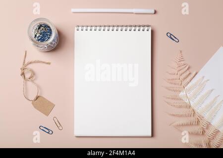 Scrivania da lavoro con notebook, penna, graffette, cartellino in carta bianca e felce su sfondo beige.Lay piatto, business, lavoro, concept mockup. Spazio per la copia. Foto Stock