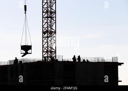 Sagome di operai e gru a torre con carico in cantiere contro il cielo. Edilizia abitativa, costruttori che lavorano su ponteggi Foto Stock