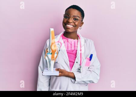 Giovane dottoressa afroamericana in possesso di modello anatomico di ginocchio giunto che guarda positivo e felice in piedi e sorridente con un spettacolo di sorriso sicuro Foto Stock